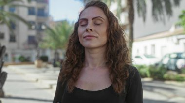 Gözleri kapalı gülümseyen bir kadın açık mavi gökyüzünün altında yüksek binalar ve yeşilliklerle çevrili bir şehir caddesinde duruyor..