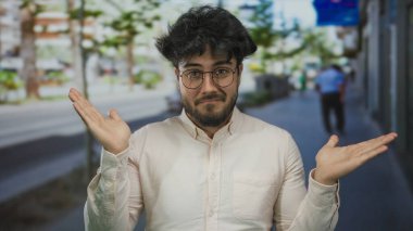 Sokakta duran sakallı ve gözlüklü genç adam bulanık şehir geçmişine karşı açık ellerle şaşkın şaşkın bakıyor..