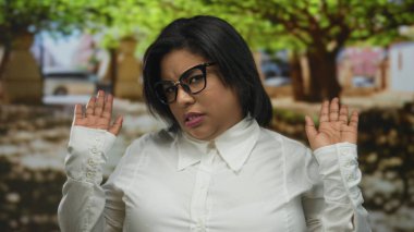 Beyaz tişörtlü bir kadın, güneşli bir ortamda umursamazlık izlenimi uyandıran ağaçlarla parkta küçümseyici jestler yapıyor..