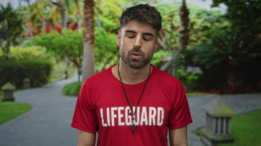 Kırmızı cankurtaran tişörtü giyen bronz tenli genç adam sokak parkında ıslıkla ayakta duruyor..