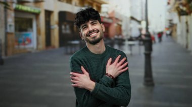 Bir kenar mahallede kendini kucaklayan genç adam, gülümseyerek kendini sevgiyle ve sükunetle ifade ediyor. Arka planda iki adam ve parlak ışık, kalabalık bir şehirde huzurlu bir anı yakalıyor.