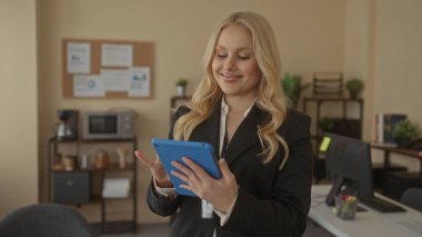 Ofiste elinde sarı saçlı ve takım elbiseli bir tablet tutan bir kadın. Etrafı ofis mobilyalarıyla çevrili, odaklanmış ve profesyonel görünüyor..