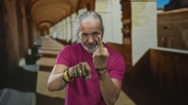 Uzun gri saçlı bir adam bina koridorunda orta parmağını gösteriyor, isyankar tavır sergiliyor..