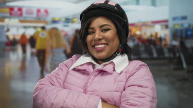 Pembe ceketli, kasklı, havaalanı terminalinde gülümseyen bir kadın güven ve pozitiflik gösteriyor..