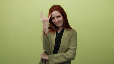 Kızıl saçlı, boynuzlu bir kadın, izole edilmiş sarı bir arka planda gülümseyen, kendine güveni ve stili olan yeşil ceketli, neşeli bir ifade sergileyen bir jest yapıyor..