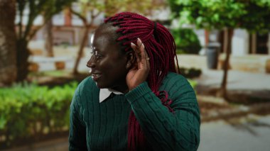 Kulaktan kulağa dikkatle dinleyen, yeşil süveter giyen, ağaçlarla çevrili ve şehir dışında yaşayan, canlı kırmızı örgüler sergileyen bir kadın..