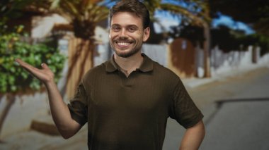 Gülümseyen bir adam, sol avuç içi yukarı ve sağ el kalça, bir ev girişinde ayakta gün ışığında; dostça karşılama.