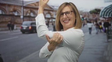 Beyaz gömlekli orta yaşlı beyaz kadın şehir caddesinde bulanık binaları ve insanları geçerek kolunu esnetiyor; güven güçlendiriyor.