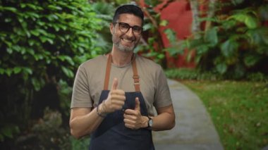 Kot önlüklü ve gözlüklü bir adam her iki işaret parmağını yeşil yapraklı bir orman yolunda kameraya doğrultuyor..