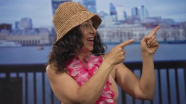 Orta yaşlı bikinili kadın, hasır şapka ve deniz kenarındaki gezinti güvertesini işaret eden lei; tatil neşesi.