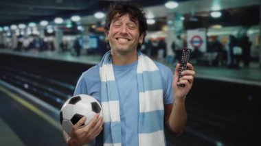 Metroda elinde futbol topu tutan İspanyol bir adam ve yeraltı ortamında uzaktan gülümseyen mavi bir eşarp takan, işe gidip gelen coşkulu bir taraftarı temsil eden.