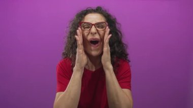 Elleri yanaklarında, ağzı mor stüdyo duvarına dayalı kırmızı tişörtlü genç İspanyol kadın. Sürpriz..