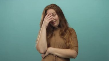 Eli yüzü örtülü genç kızıl saçlı kadın stüdyoda donuk arkaplan ile kollarını kavuşturuyor; utanç..