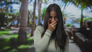 Parkta genç bir kadın, yemyeşil bir yeşillik ve parlak mavi gökyüzünün altında palmiyelerle çevrili, neşeli bir gülümsemeyle, açık havayı kucaklıyor..