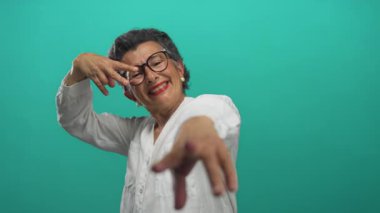 Gri saçlı yaşlı kadın yeşil arka planda barış jesti yapıyor. Gözlük takıyor ve beyaz bir gömlek giyiyor. Canlı bir ortamda neşe ve pozitiflik gösteriyor..