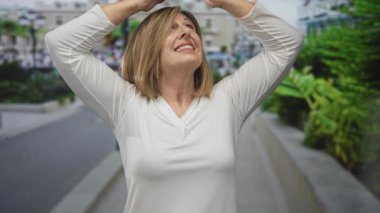 Beyaz gömlekli orta yaşlı kadın, parlak güneş ışığı altında bir şehir sokağında elleri olan gözleri gölgeliyor; iyimserlik.