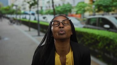 Kıvırcık saçlı ve gözlüklü gülümseyen kadın, şehir sokağında elleri açık, canlı sarı gömlekli, neşe ve pozitiflik saçan bir kalp yapar..