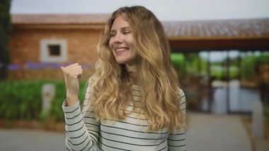 Kadın bir zafer sahnesinde elini kaldırır. Sarışın genç bir sanatçıyla bir bahçe sahnesinin ortasında. Geniş gülümsemesi açık havada bir evin önünde dalgalanan saçlarını ön plana çıkarır..