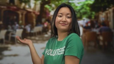 Yeşil tişörtlü bir kadın, arka planda kahvelerini yudumlayan insanların olduğu bir teras restoranında jest yapıyor..