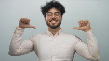 Sakallı, gözlüklü, beyaz gömlekli, beyaz arka planda sırıtan genç bir adam iki başparmağını da kaldırmış kendini işaret ediyor..