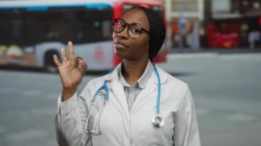 Şehir merkezindeki kadın doktor, kameraya güvenle gülümseyen beyaz önlüklü ve gözlüklü, steteskoplu iyi bir jest yapıyor.