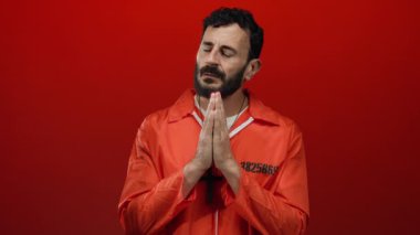 Turuncu hapishane tulumu giymiş İspanyol bir adam elleri dua ederken kırmızı arka planda duruyor..