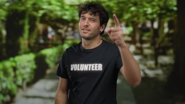 Gönüllü tişört giyen İspanyol bir adam yeşil bir parkta duruyor, kamerayı işaret ediyor, yemyeşil ağaçlar ve güneşli yollarla çevrili..