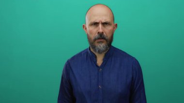 Yeşil arka planda ciddi bir ifadeye sahip mavi gömlekli bir adam kararlı bir görünüm sergiliyor.