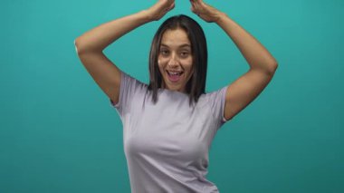 Latin kökenli genç bir kadın kollarını başının üstüne kaldırıyor ve stüdyosunda tebessüm ve lavanta rengi t-shirt ile gülümsüyor..