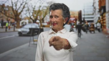 Gri saçlı yaşlı kadın gülümsüyor ve şehir arka planında bulanık bir şehir sokağındaki kameraya bakıyor, hareketli bir kasabada canlı bir açık hava manzarası yakalıyor..