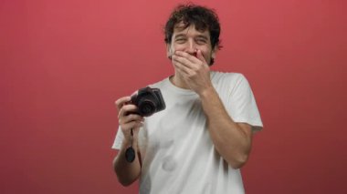 İspanyol bir adam elinde kamerayla kırmızı bir zemine gülüyor, neşeli bir mizah ve yaratıcılık anı yakalıyor..