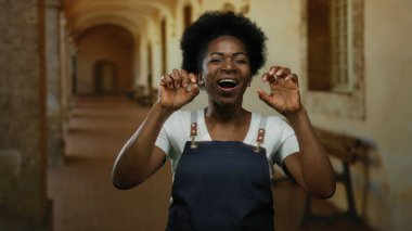 Önlüklü kadın, tarihi üniversite koridorunda klasik bir ortamda neşe, kültürel çeşitlilik ve eğitim sergileyerek kapalı mekan jestleri yapıyor..