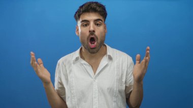 Genç, ağzı açık bir İspanyol adam, parlak mavi stüdyoda ellerini kaldırdı ve şaşırmış ifade; sürpriz şaşkınlık.