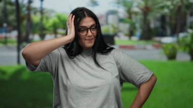 Parkta gözlüklü bir kadın poz veriyor, büyük beden bir figür sergiliyor, yeşillik ve şehir zemininin ortasında duruyor, duygularını ifade ediyor, uzun siyah saçlar ve günlük gri bluzuyla..