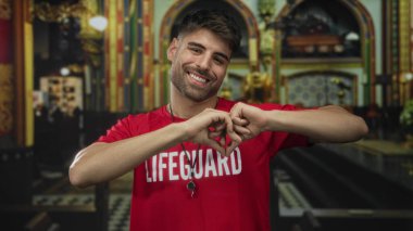 Kırmızı cankurtaran gömleği giyen adam elleriyle kalp şekli oluşturuyor ve kilisenin içinde geniş bir gülümseme sergiliyor; şefkat..