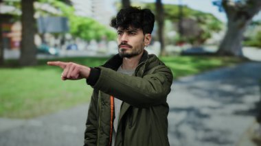 Sakallı genç adam, arka planda kararlılık ve odaklanma gösteren ağaçlar ve binalarla dolu bir parkta parkı gösteren bir ceket giyiyor..