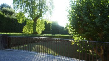 Yumuşak bokeh ve çözünmüş demir korkuluklu bulanık sahne güneşli ağaçların altında bir bahçede, sığ odak dışarıda; arka plan telif alanı sakin.