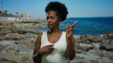 Mavi gökyüzünün altındaki kayalık plajda el kol hareketi yapan bir kadın, doğal deniz manzarası sergiliyor, ifadesini ve sahil ortamında dış görünüşünü vurguluyor..