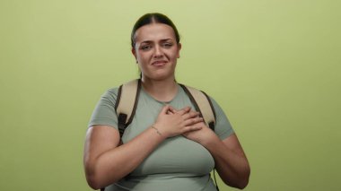 Sırt çantasıyla göğsünü tutan kıvrımlı genç bir kadın izole edilmiş sarı bir duvarın karşısında duruyor. Rahatsızlık ve endişeyi ifade ediyor. Duygularını ve gerginliğini ifade ediyor..