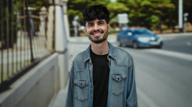 Şehirdeki bir sokakta gülümseyen genç bir adam. Sıradan bir kot ceket giyiyor. Kendinden emin ve rahat görünüyor..