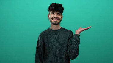 Genç bir adam yeşil bir arka planda gülümseyerek avucunun içini işaret ediyor, kendine güveni ve cazibesi ile süveter giymiş sıradan bir stile sahip..