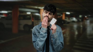 Kot ceketli genç adam loş ışıklı bir park alanında bir kart tutuyor, dikkatlice bakıyor, tefekkür etmeyi ya da karar vermeyi öneriyor..