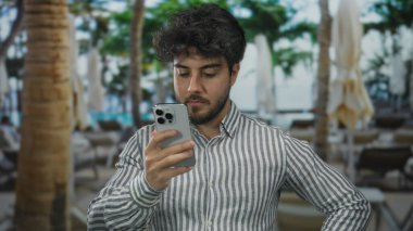 Sakallı genç adam arka planda palmiye ağaçları ve tropikal atmosferi olan açık hava restoranının terasında akıllı telefon kullanarak iletişim için rahat bir atmosfer yaratıyor.