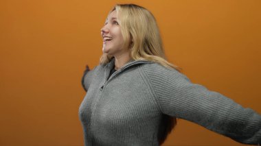 Turuncu bir duvarın önünde kollarını açmış gülümseyen, rahat gri bir kazak giyen, neşe ve özgürlük duygusu taşıyan bir kadın..