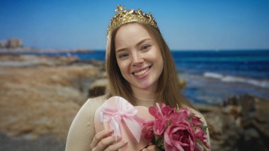 Taç giyen genç kadın açık gökyüzünün altında kumlu bir sahil şeridinde kalp şeklinde hediye kutusu ve şakayık buketi tutuyor..