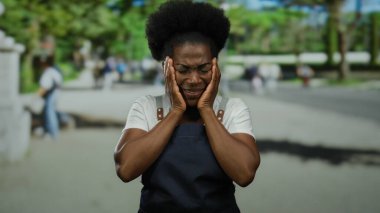 Açık şehir caddesinde elleri açık önlük giyen bir kadın, bulanık arka plan ve yeşil yeşillikler arasında duygu ve içgözlem taşıyor..