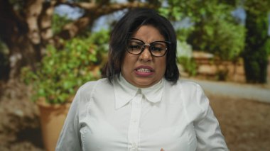 Parkta ağzı, gözlüğü ve beyaz gömleği olan, yeşilliklerle çevrili, açık hava yaz ortamında duyguları somutlaştıran bir kadın iğreniyor..