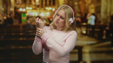 Katolik kilisesinde bilek ağrısı çeken, kulaklık takan, dingin ortamın ortasında endişeli görünen, modern ruhani bir anı yakalayan bir kadın..