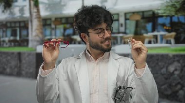 Güneşli bir günde, dışarıda gözlük tutan sakallı genç bir adam. Parlak güneş ışığına hangi çerçevenin daha iyi uyacağını seçerken yüzünde bir endişe ifadesi var.