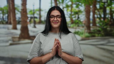 Uzun siyah saçlı, uzun boylu, gözlüklü ve gri gömlekli, palmiye ağaçları ve bulanık yeşilliklerle çevrili genç bir kadın..